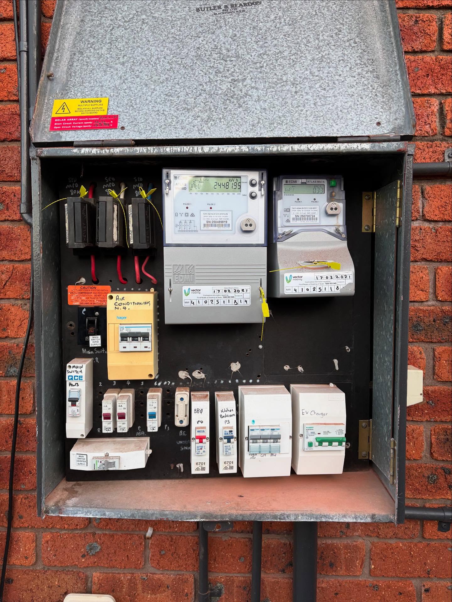 an older switchboard in a galvanised meter box on a red brick wall, with a mix of old fuses and newer circuit breakers